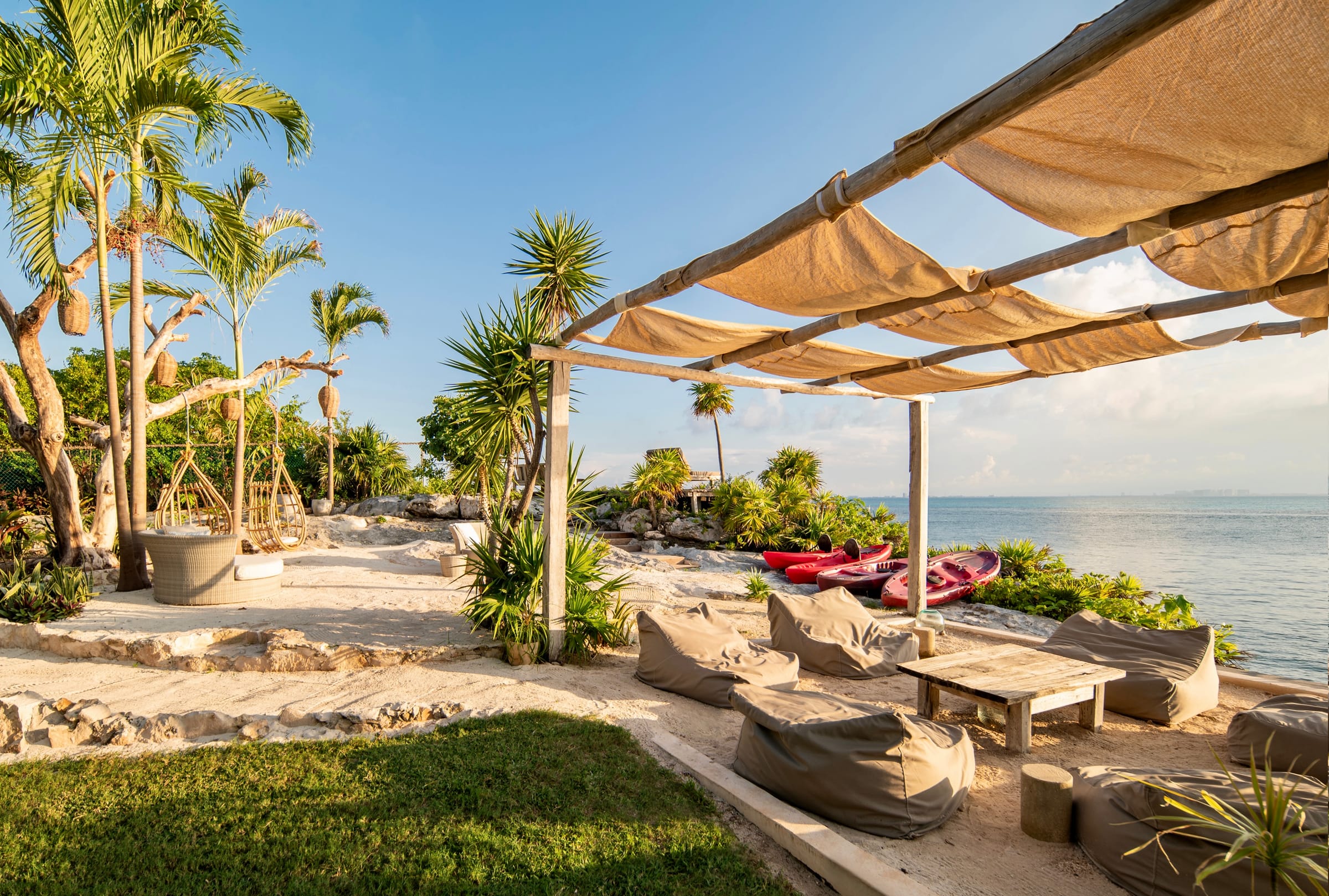 Shaded canvas pergola over beach seating at the water's edge, kayaks waiting beyond