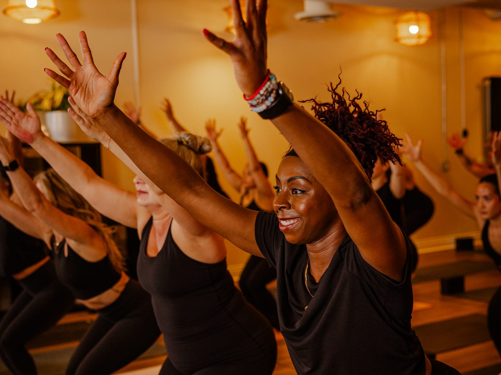 A smiling member reaching overhead in chair pose during a heated class at The Lineage